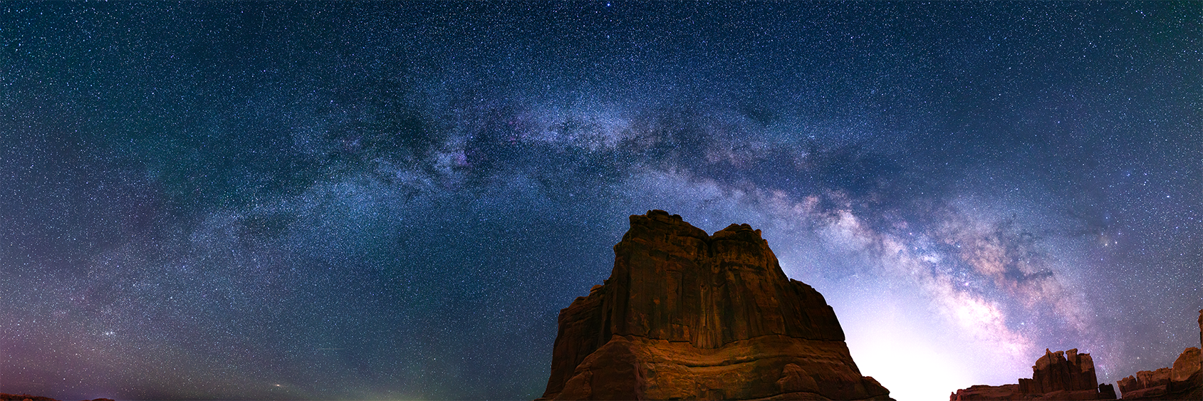 The Monolith Smiles Milky Way galaxy glowing above dramatic sandstone towers at night in Arches National Park, Utah